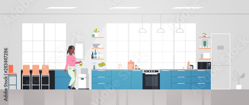 african american woman putting trash in electronic recycling bin controlled by smart speaker voice recognition activated digital assistant concept modern kitchen interior horizontal full length