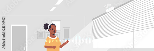 african american woman using smart speaker voice recognition activated digital assistant concept modern living room interior flat horizontal portrait