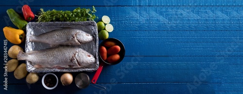 Preparando Peixe Corvina Brasileira com Pimentão Vermelho, Pimentão Verde, Pimentão Amarelo, Salsinha, Batata, Azeitona Preta, Cebola, Tomates Italianos e limão sobre a mesa de madeira azul marinho photo