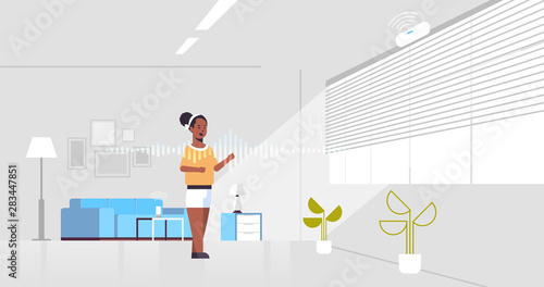 african american woman using smart speaker voice recognition activated digital assistant concept modern living room interior flat horizontal full length