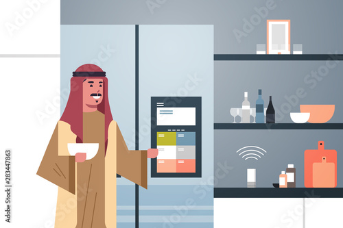 arab man touching refrigerator screen with smart speaker voice recognition activated digital assistant concept modern kitchen interior flat horizontal portrait