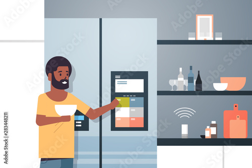 african american man touching refrigerator screen with smart speaker voice recognition activated digital assistant concept modern kitchen interior flat horizontal portrait