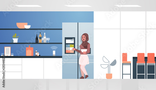 arab woman touching refrigerator screen with smart speaker voice recognition activated digital assistant concept modern kitchen interior flat horizontal full length