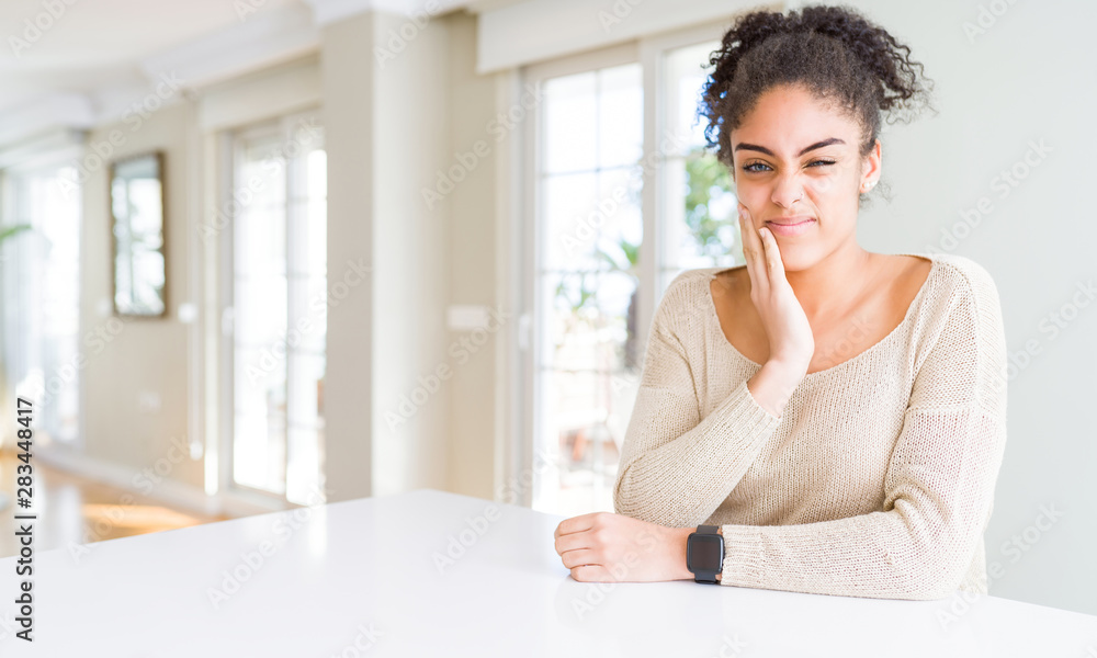 Beautiful young african american woman with afro hair sitting on table at home touching mouth with hand with painful expression because of toothache or dental illness on teeth. Dentist concept. Beautiful young african american woman with afro hair sitting on table at home touching mouth with hand with painful expression because of toothache or dental illness on teeth. Dentist concept.