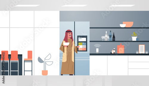 arab man touching refrigerator screen with smart speaker voice recognition activated digital assistant concept modern kitchen interior flat horizontal full length