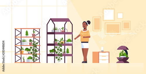 african american woman using automatic watering system controlled by smart speaker voice recognition activated assistant concept modern smart home garden interior flat full length horizontal