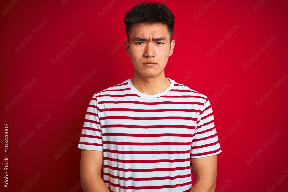 Young asian chinese man wearing striped t-shirt standing over isolated red background skeptic and nervous, frowning upset because of problem. Negative person. Young asian chinese man wearing striped t-shirt standing over isolated red background skeptic and nervous, frowning upset because of problem. Negative person.