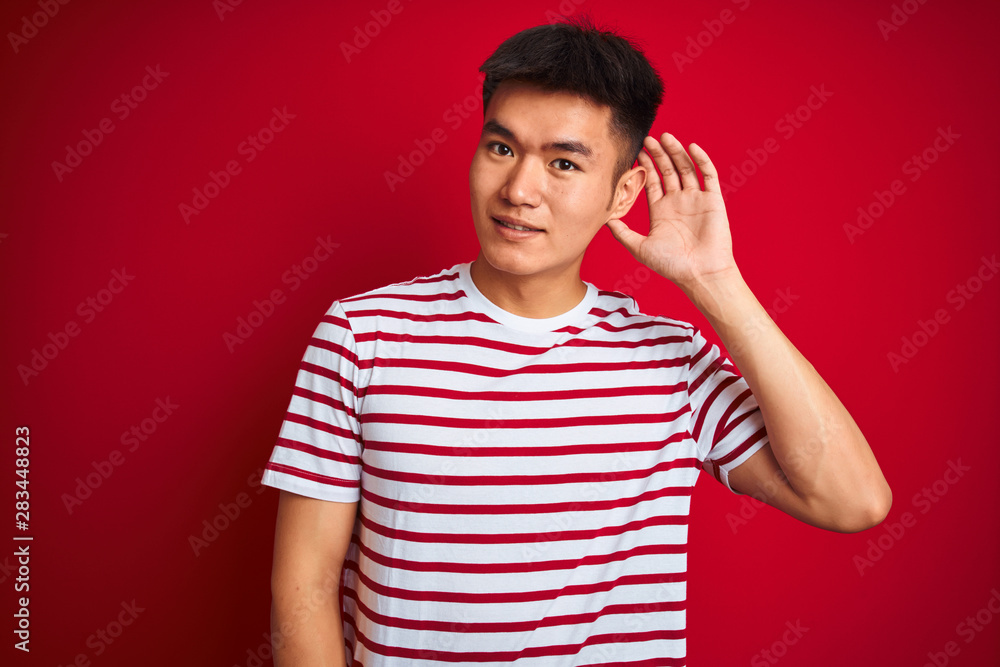 Young asian chinese man wearing striped t-shirt standing over isolated red background smiling with hand over ear listening an hearing to rumor or gossip. Deafness concept. Young asian chinese man wearing striped t-shirt standing over isolated red background smiling with hand over ear listening an hearing to rumor or gossip. Deafness concept.