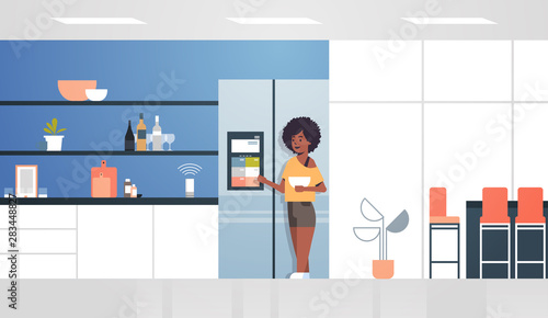 african american woman touching refrigerator screen with smart speaker voice recognition activated digital assistant concept modern kitchen interior flat horizontal full length