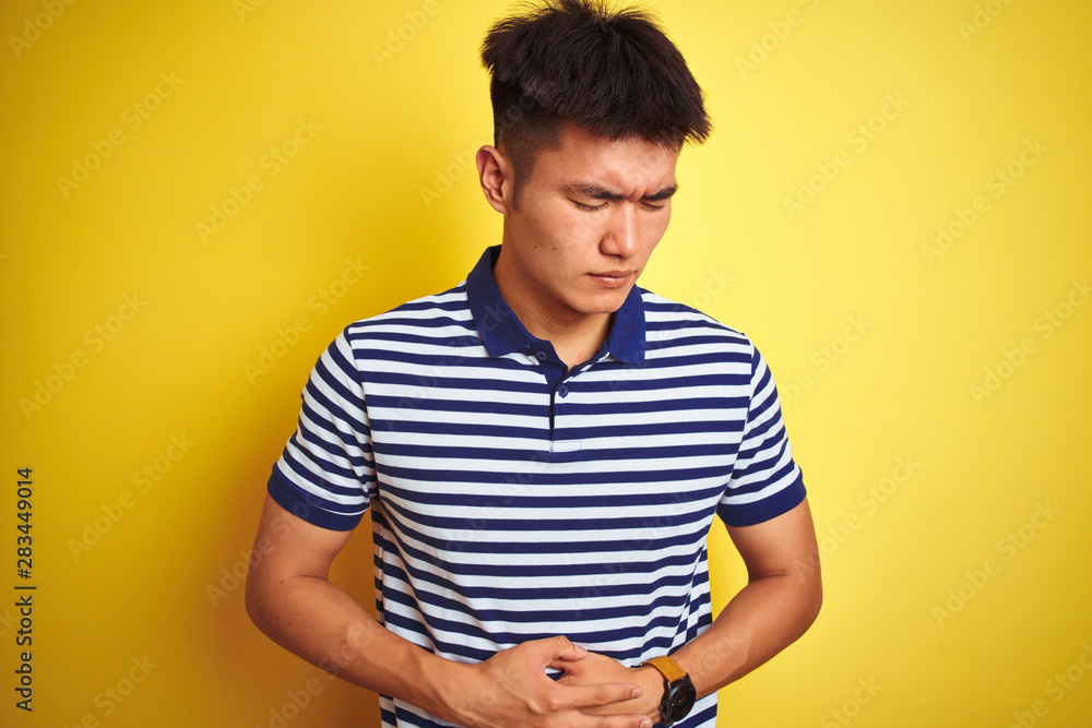 Young asian chinese man wearing striped polo standing over isolated yellow background with hand on stomach because nausea, painful disease feeling unwell. Ache concept. Young asian chinese man wearing striped polo standing over isolated yellow background with hand on stomach because nausea, painful disease feeling unwell. Ache concept.