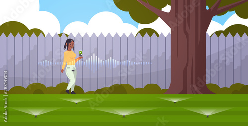 african american woman using mobile app controlling irrigation grass watering system by smart speaker voice recognition activated assistant concept backyard with green lawn horizontal full length
