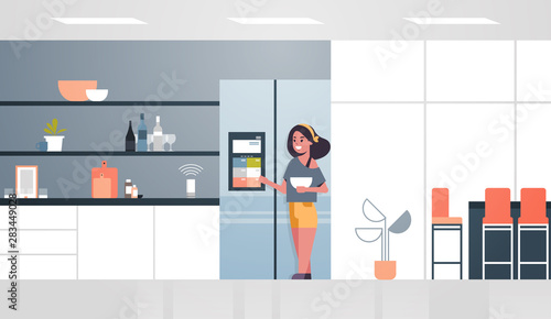 woman touching refrigerator screen with smart speaker voice recognition activated digital assistant concept modern kitchen interior flat horizontal full length © mast3r