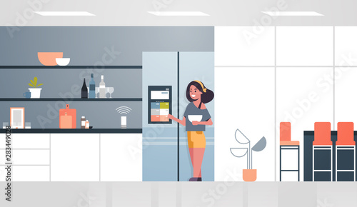 woman touching refrigerator screen with smart speaker voice recognition activated digital assistant concept modern kitchen interior flat horizontal full length
