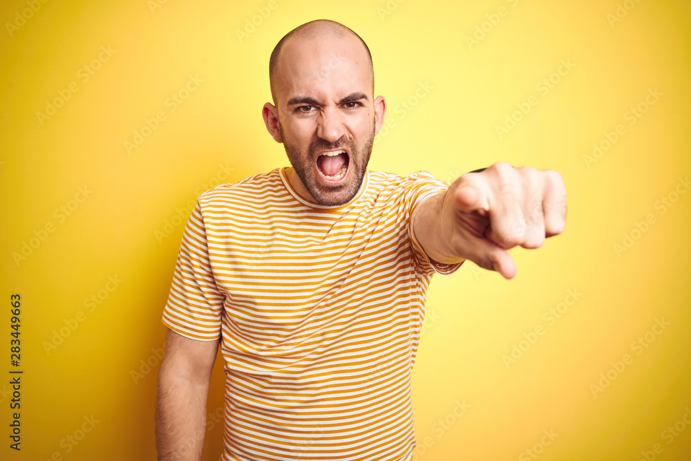Young bald man with beard wearing casual striped t-shirt over yellow isolated background pointing displeased and frustrated to the camera, angry and furious with you Young bald man with beard wearing casual striped t-shirt over yellow isolated background pointing displeased and frustrated to the camera, angry and furious with you