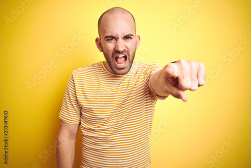 Young bald man with beard wearing casual striped t-shirt over yellow isolated background pointing displeased and frustrated to the camera, angry and furious with you