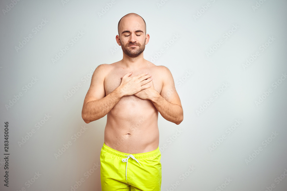 Young shirtless man on vacation wearing yellow swimwear over isolated background smiling with hands on chest with closed eyes and grateful gesture on face. Health concept.