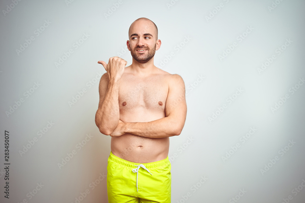 Young shirtless man on vacation wearing yellow swimwear over isolated background smiling with happy face looking and pointing to the side with thumb up.
