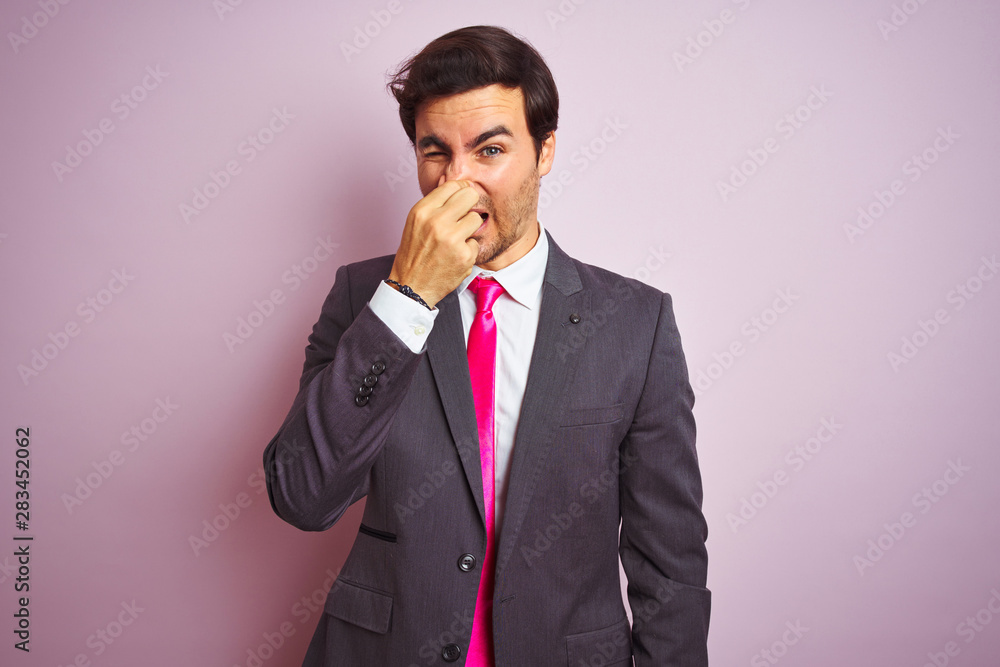 Young handsome businessman wearing suit and tie standing over isolated pink background smelling something stinky and disgusting, intolerable smell, holding breath with fingers on nose. Bad smells Young handsome businessman wearing suit and tie standing over isolated pink background smelling something stinky and disgusting, intolerable smell, holding breath with fingers on nose. Bad smells