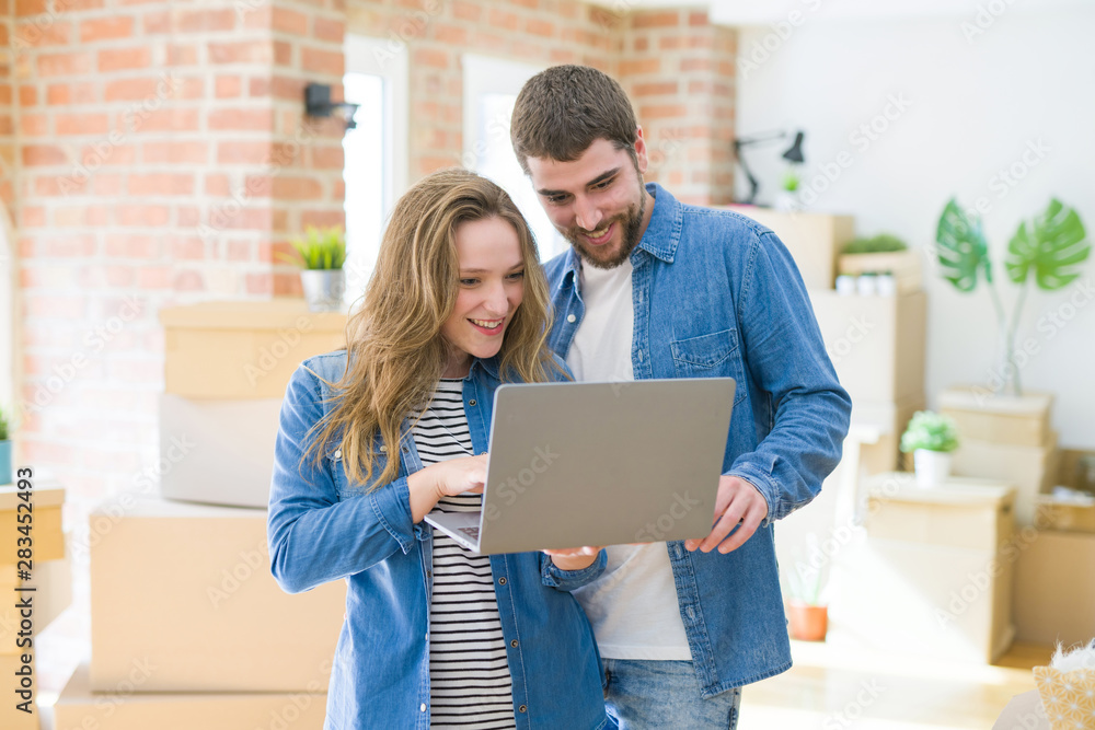 Young couple using computer laptop standing on a room around cardboard boxes, happy for moving to a new apartment Young couple using computer laptop standing on a room around cardboard boxes, happy for moving to a new apartment