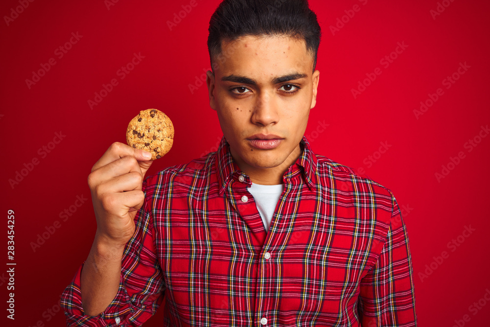 Young brazilian man holding a cookie standing over isolated red background with a confident expression on smart face thinking serious Young brazilian man holding a cookie standing over isolated red background with a confident expression on smart face thinking serious
