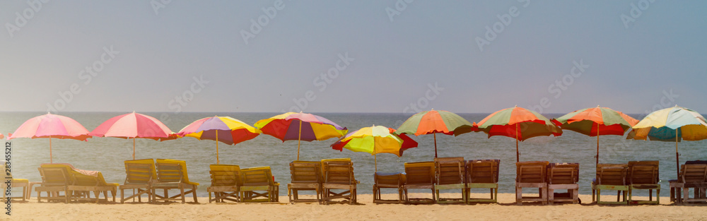 Horisontal banner - row of empty wooden chaise lounges with colorful umbrellas on the beach in GOA, India, toned with sunlight. Sea summer holiday background.