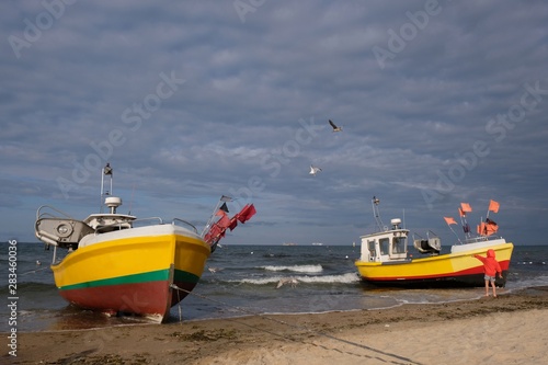 Two wooden fishing boats on the beach of Baltic Sea in Sopot/Poland. Silhouette of little boy in red jacket standing near boat and pointing his hand at flying seagulls.