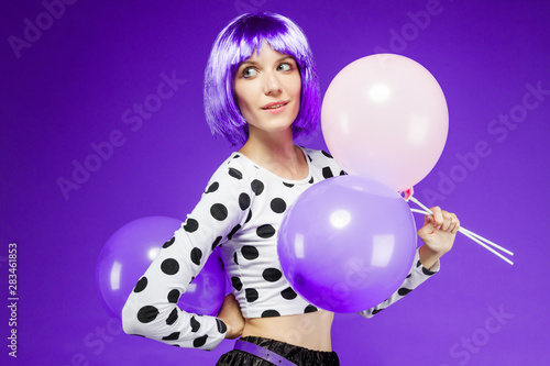 Beautiful thoughtful girl in wig is holding pink and violet balloons. Stylish fashion woman dressed in trends of season. Bright young female on purple background. Good mood and happy lifestyle concept