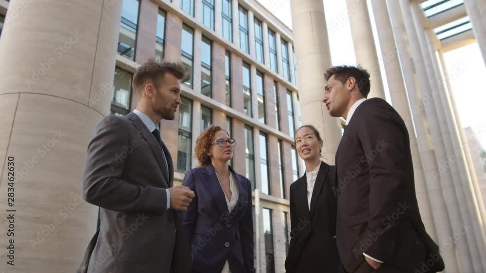 Low angle view of two businessmen and their two female colleagues standing together on street and chatting Low angle view of two businessmen and their two female colleagues standing together on street and chatting