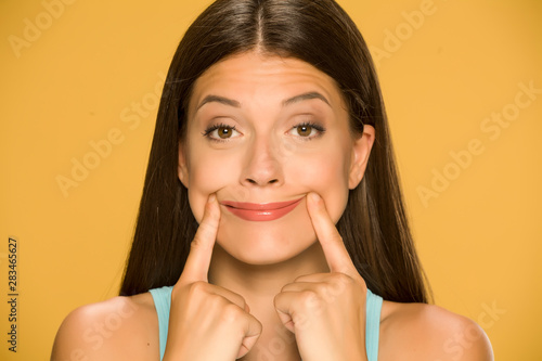 Young woman forcing her smile with her fingers on yellow background
