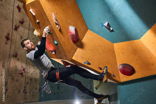Full length shot of physically impaired male mountaineer practicing rock climbing at indoor bouldering settings, dressed in comfortable sport clothes, equipped with harness and chalk bag.