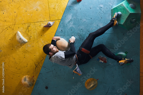 Side view of motivated active physically challenged boulderer with beard hanging at colourful climbing wall, holding large artificial rock, trying to fix his leg at feet hold. Indoor sport concept.