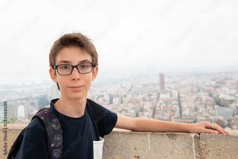 Handsome young boy wearing glasses posing over background of panorama of Alicante (Spain). City view from Mount Santa Barbara with teen foreground