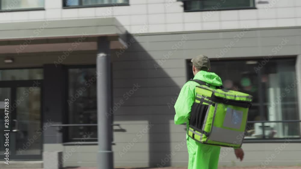 Tracking shot of young male food courier walking down street with insulated backpack using cell phone for navigation. Food delivery guy coming to house door, calling doorbell and speaking to customer Tracking shot of young male food courier walking down street with insulated backpack using cell phone for navigation. Food delivery guy coming to house door, calling doorbell and speaking to customer