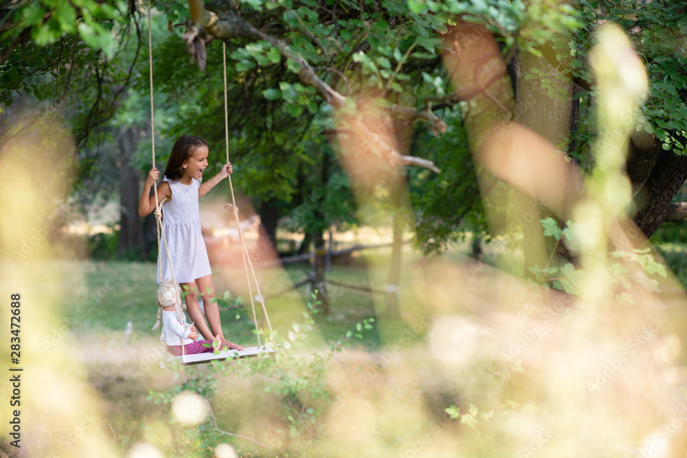 Happy girl rides on a rope swing with her retro doll in park. Little Princess has fun outdoor, summer nature outdoor. Childhood, child lifestyle, enjoyment, happiness.