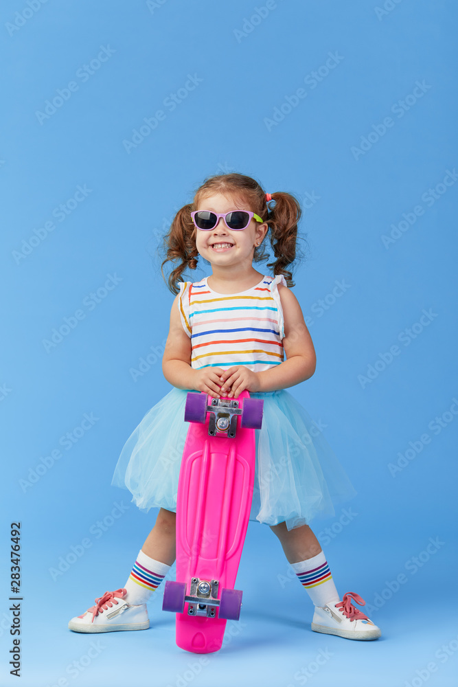 Stylish little toddler girl child in glasses with skateboard over blue background Stylish little toddler girl child in glasses with skateboard over blue background