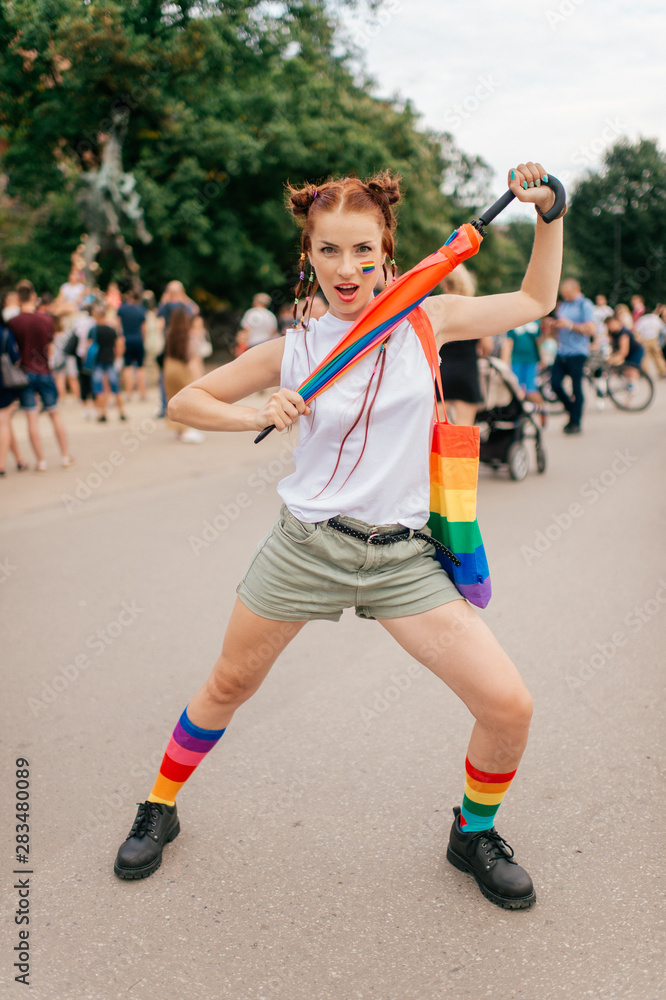 bisexual girl with lgbt gay pride flag on her face, colorful umbrella, bag and mismatched socks posing on street with people on background.