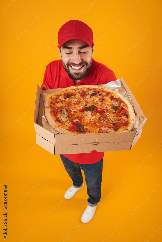 Full length image of optimistic delivery man in red uniform smiling and holding pizza box