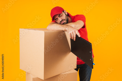 Image of young delivery man in red uniform sleeping while standing with clipboard over packaging boxes