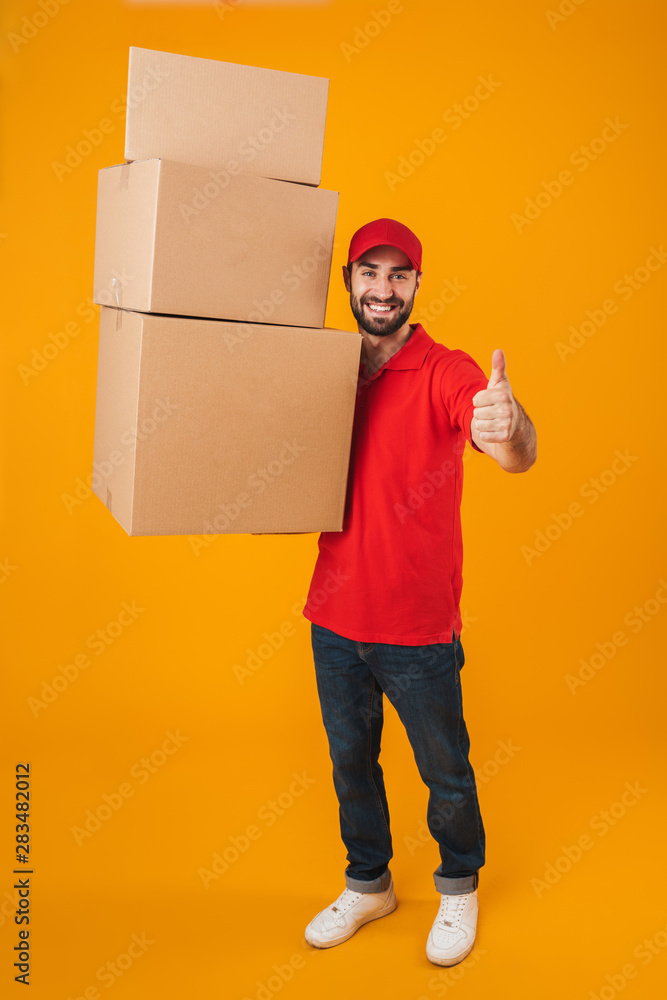 Full length portrait of handsome delivery man in red uniform showing thumb up while carrying packaging boxes - obrazy, fototapety, plakaty 