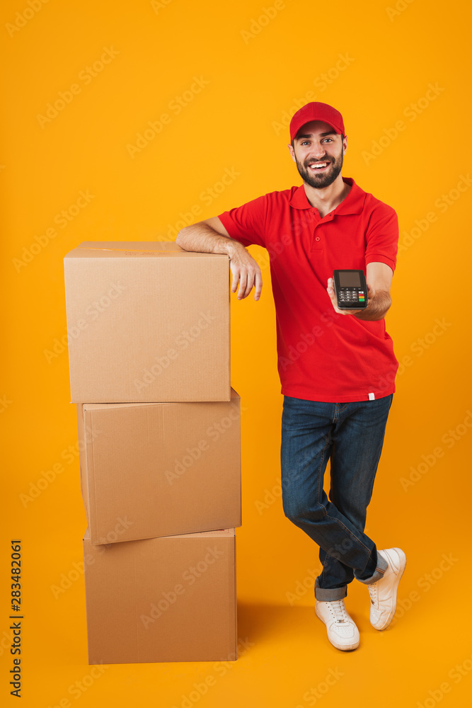 Portrait of caucasian delivery man in red uniform holding payment terminal while standing with packaging boxes Portrait of caucasian delivery man in red uniform holding payment terminal while standing with packaging boxes