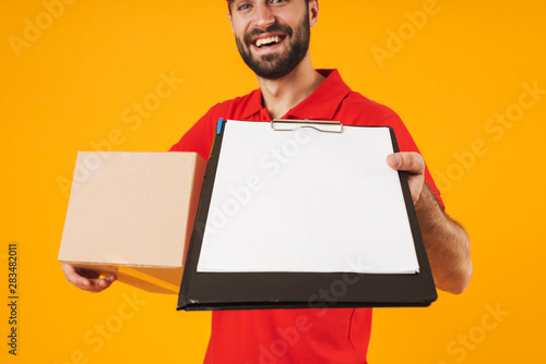 Image of handsome delivery man in red uniform holding clipboard and carrying packaging box