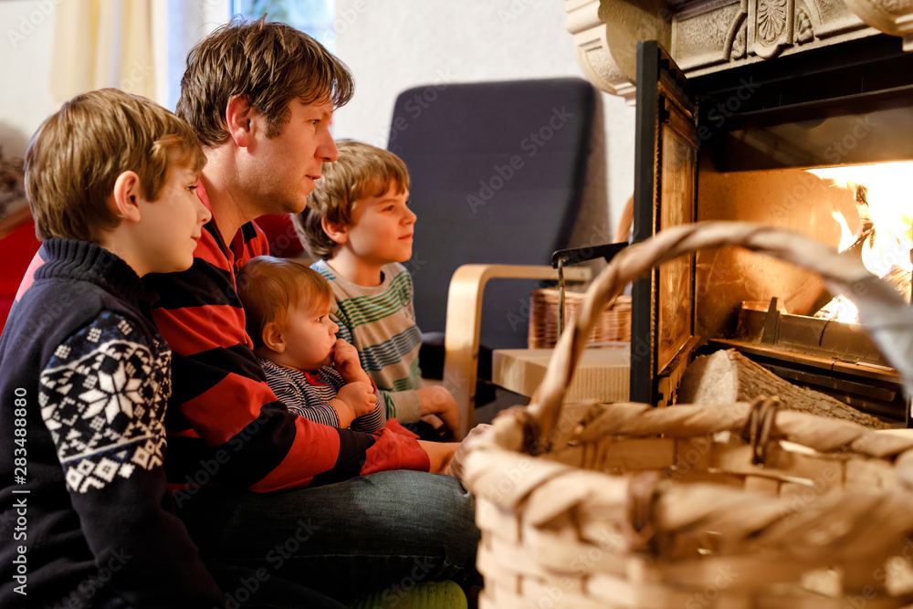 Middle age father sitting by fireplace with cute little baby girl and two kids boys at home. Happy family, dad with daughter and sons make fire for Christmas time. Safe handling for kids