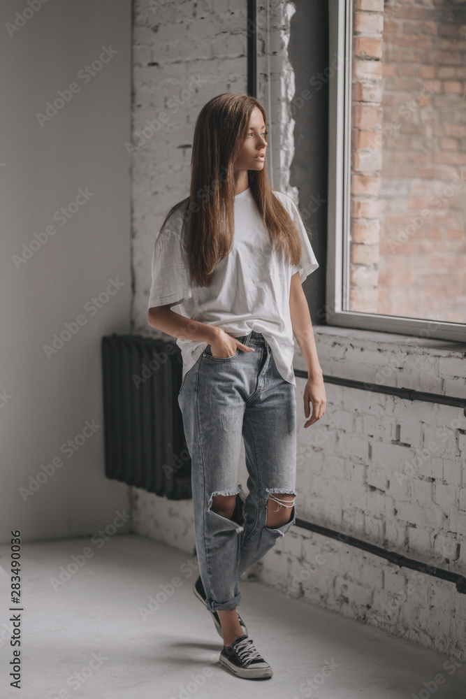 Seductive portrait of young sexy girl with brunette hair in shirt, jeans  and sneakers on white background in room near window Stock Photo | Adobe  Stock