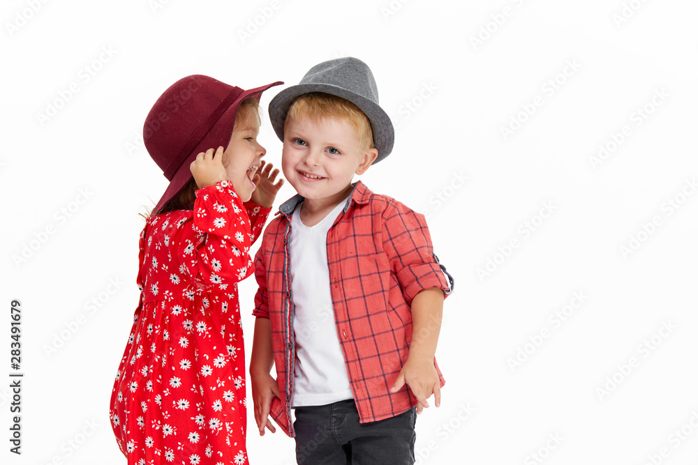 Happy brother and sister in hats embracing each other. Little toddler girl whispers in the boy’s ear and hugs him on white background. Autumn clothes Happy brother and sister in hats embracing each other. Little toddler girl whispers in the boy’s ear and hugs him on white background. Autumn clothes