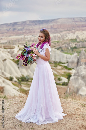 Woman with a beautiful bouquet of flowers in her hands stands on the mountain in the rays of the dawn sunset. Beautiful white long dress on the girl body. Perfect bride with pink hair