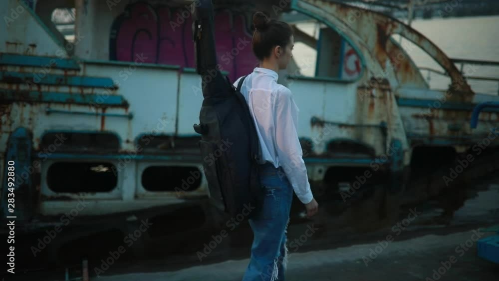 The girl in white shirt and ripped jeans stands next to old abandoned ship, puts a case with guitar on his back and leaves. The girl in white shirt and ripped jeans stands next to old abandoned ship, puts a case with guitar on his back and leaves.