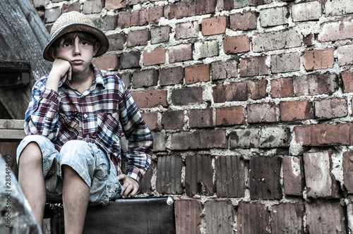 Poor sad boy sits on a suitcase against a brick wall. Homeless lonely child