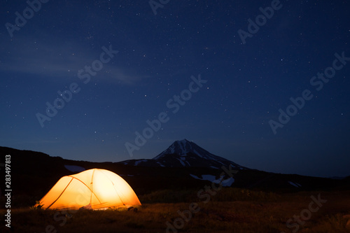 Night photo of the camping site in Kamchatka, Russia. Yellow tent in the darkness. Stars in the sky. Silhouette of smmetrical cone of Vilyuchik volcano in dark blue background.