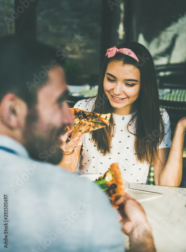 Young happy caucasian couple sitting at table eating pizza in Italian cuisine cafe on sunny summer day