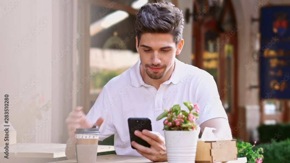 Young happy handsome man sitting in cafe outdoors at the street using mobile phone drinking coffee Young happy handsome man sitting in cafe outdoors at the street using mobile phone drinking coffee